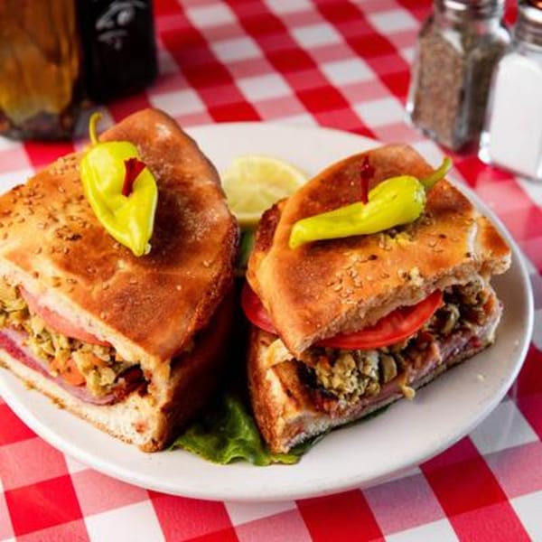 Two halves of a Monjunis Italian muffuletta sandwich with peppers on a white plate and red checkered tablecloth.